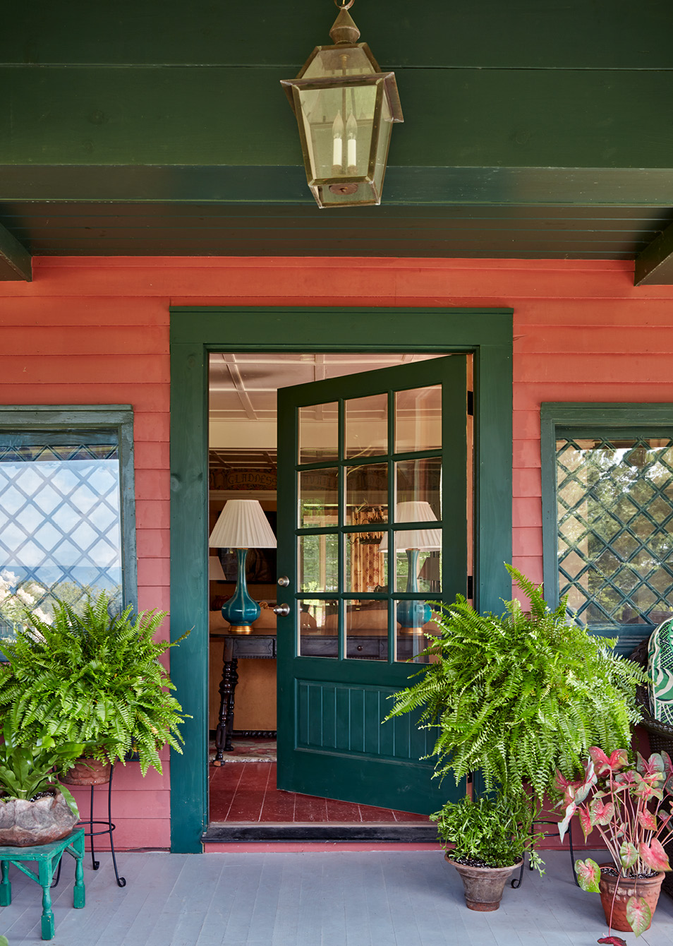 If so, you’re in for a treat today. Todays historic Catskill cottage tour designed by the talented team at Amanda Reynal Interiors is a refuge from the fast pace of modern life.   Charming and livable with lush surroundings and even lusher interiors, this home will make you feel a world away without ever leaving your living room. Thomas Loof Photography - green and red cottage with lantern hanging on front porch
