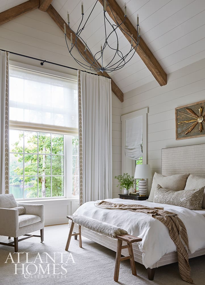 Neutral primary suite bedroom featuring wood beams, shiplap walls, and a narrow wood bench at the foot of the bed with a scenic water view