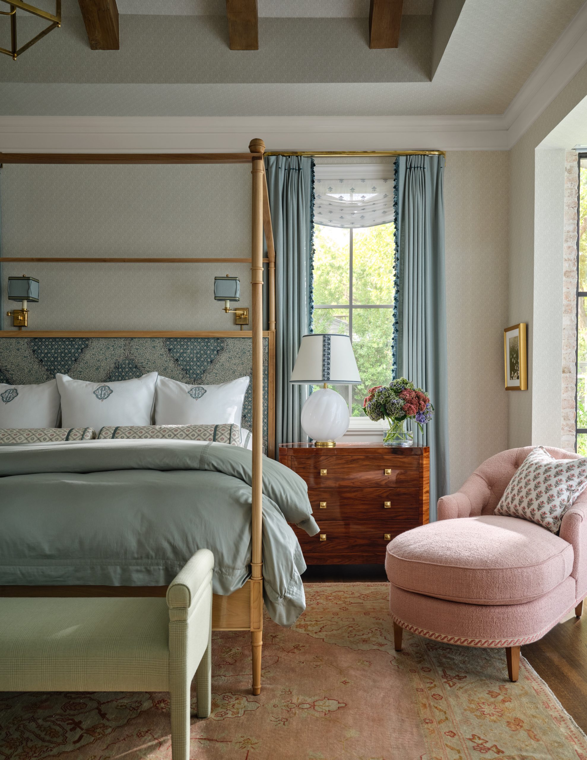 Master bedroom with a wooden canopy bed, blue upholstered headboard, green duvet, light blue curtains, a burl wood nightstand, and a pink chaise lounge chair designed by Collins Interiors.