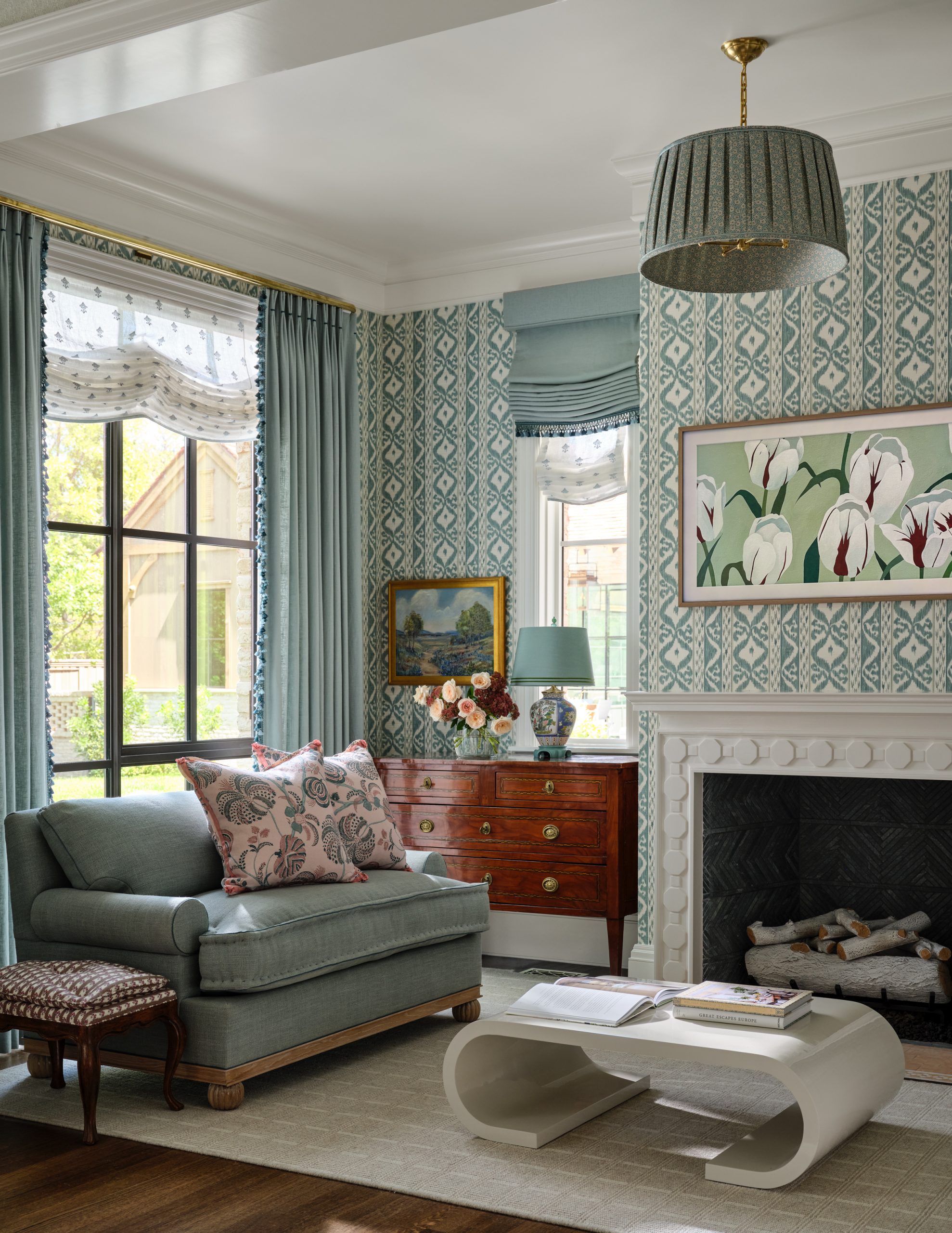 Sitting room featuring blue geometric wallpaper, a light blue armchair with floral pillows, blue tasseled curtains, a wooden dresser, a modern white coffee table, and a fireplace designed by Collins Interiors.