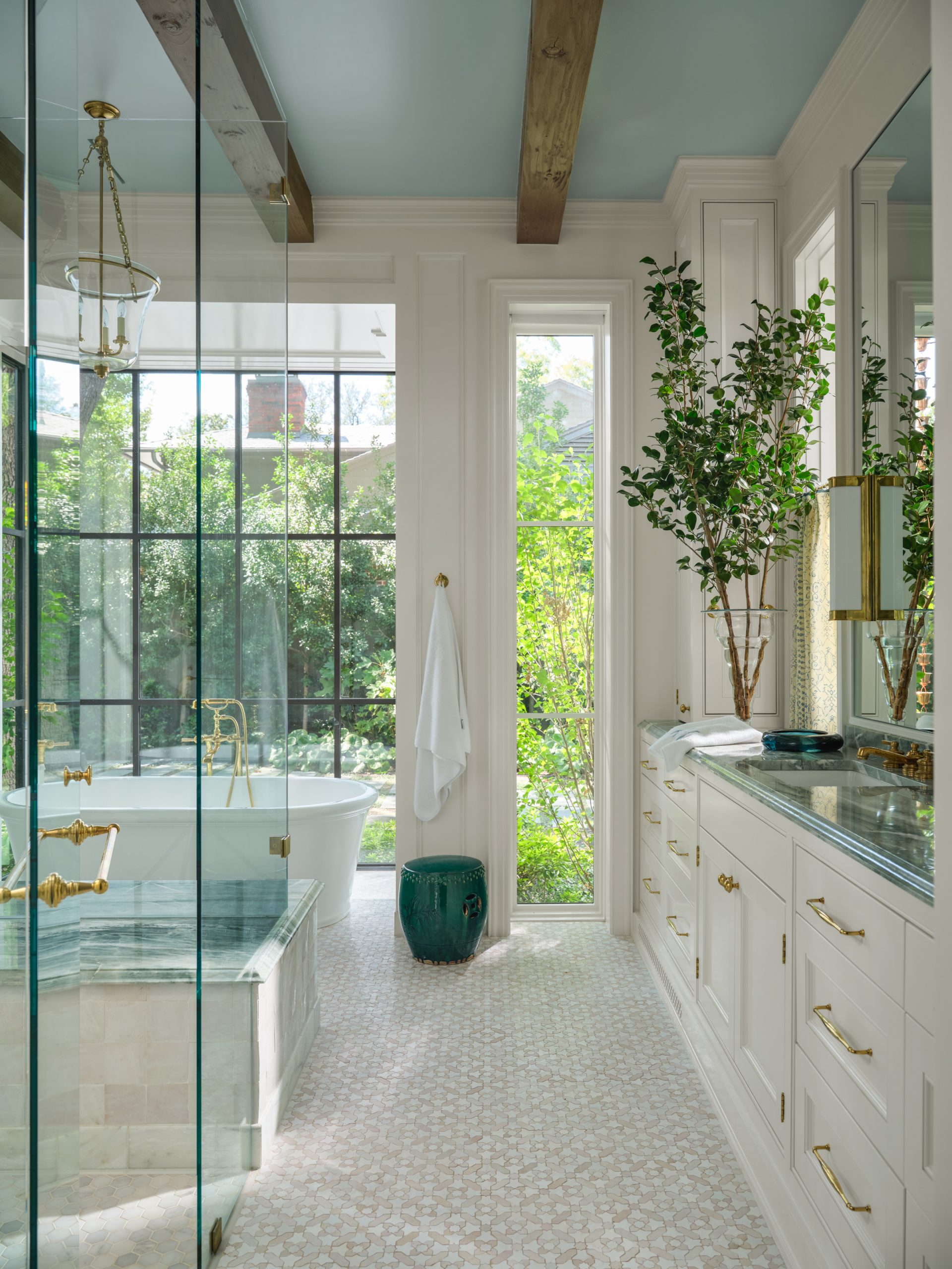 Luxury bathroom with a freestanding soaking tub, glass walk-in shower with green marble trim, white vanity with gold hardware, mosaic tile flooring, and a blue painted ceiling with wood beams designed by Collins Interiors.