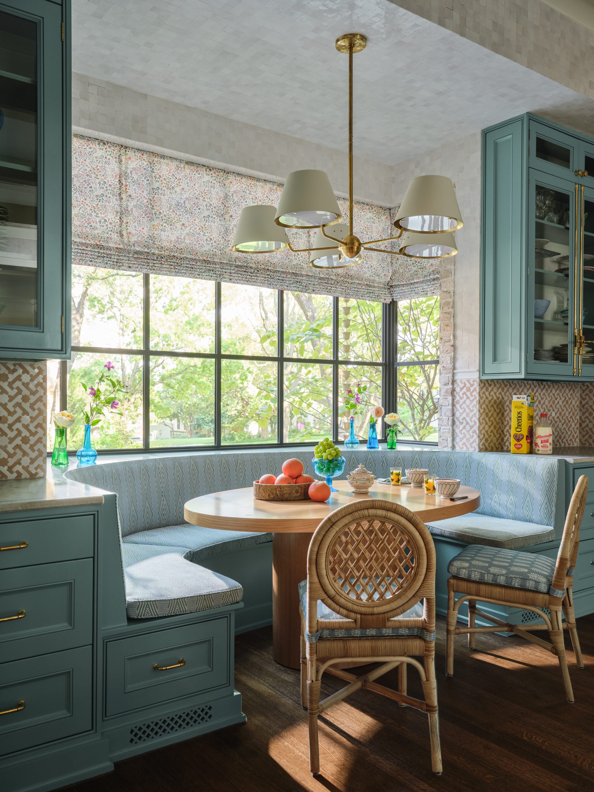 Kitchen dining nook featuring a built-in blue banquette bench, round wooden pedestal table, woven rattan dining chairs, blue cabinets with brass hardware, and a brass chandelier designed by Collins Interiors.