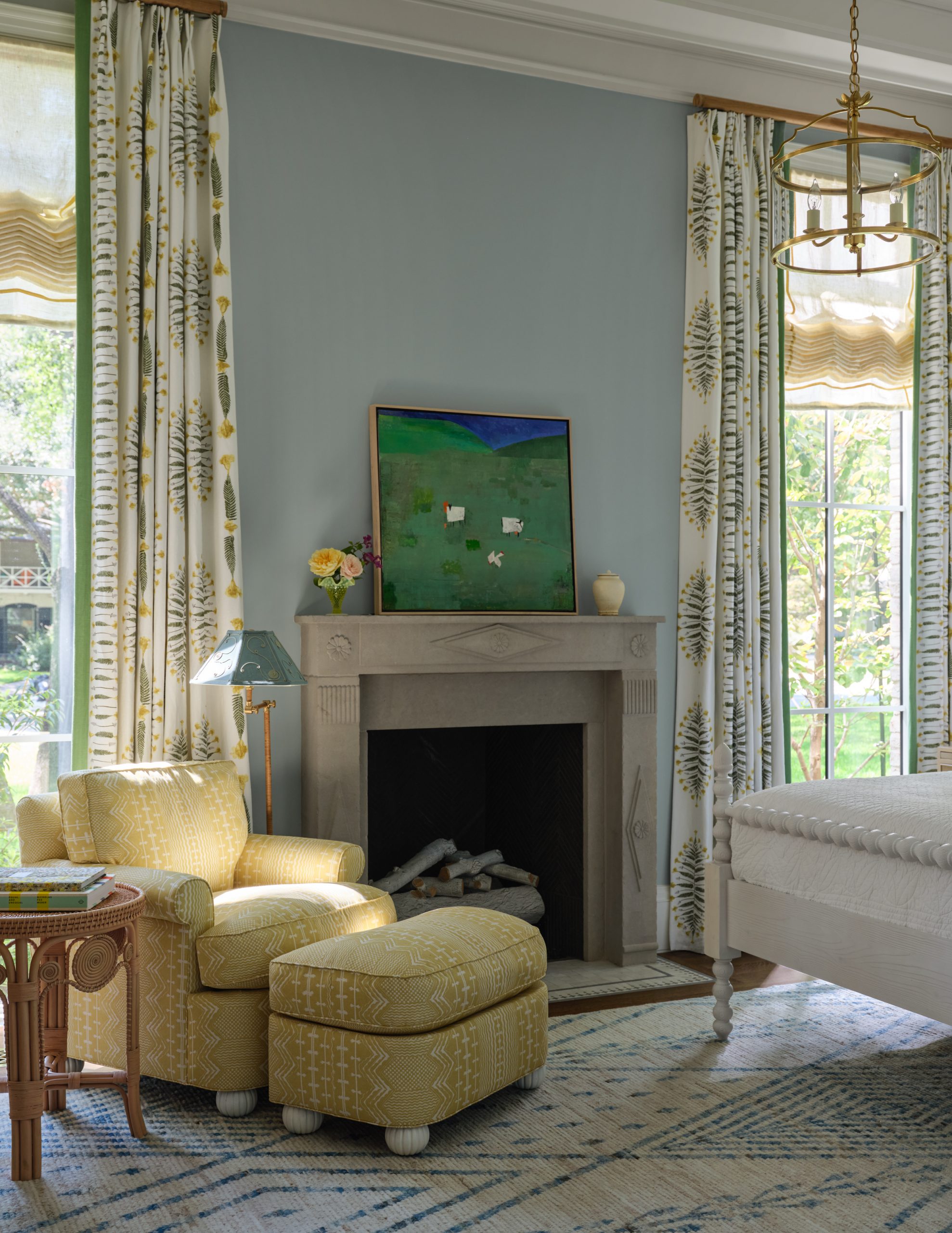 Bedroom seating area featuring a light grey stone fireplace mantel, yellow patterned armchair with a matching ottoman, light blue walls, white curtains with a yellow botanical pattern, and a green landscape painting designed by Collins Interiors.