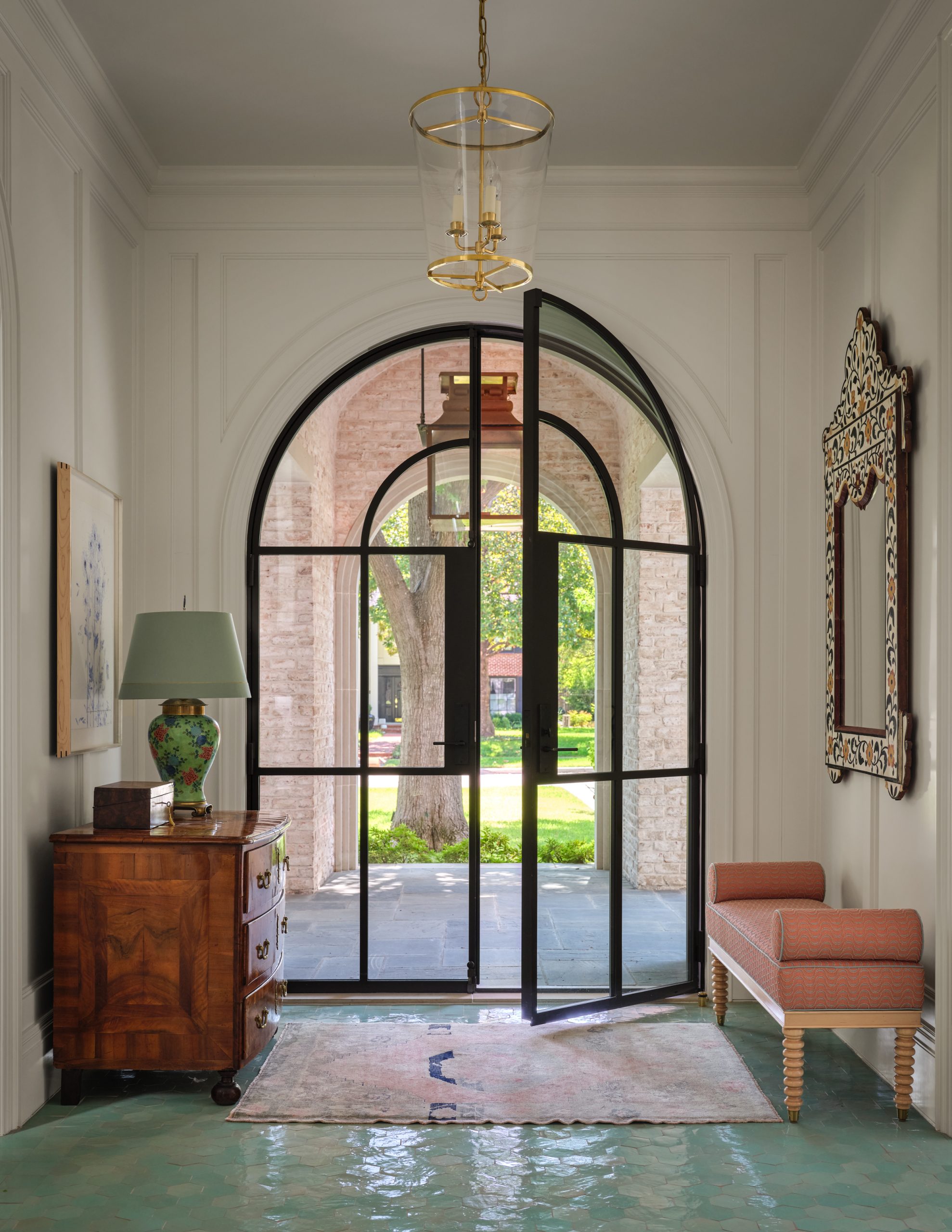 Entry foyer with a black steel arched front door, glossy green tile flooring, a vintage wood chest, and a coral bench designed by Collins Interiors.