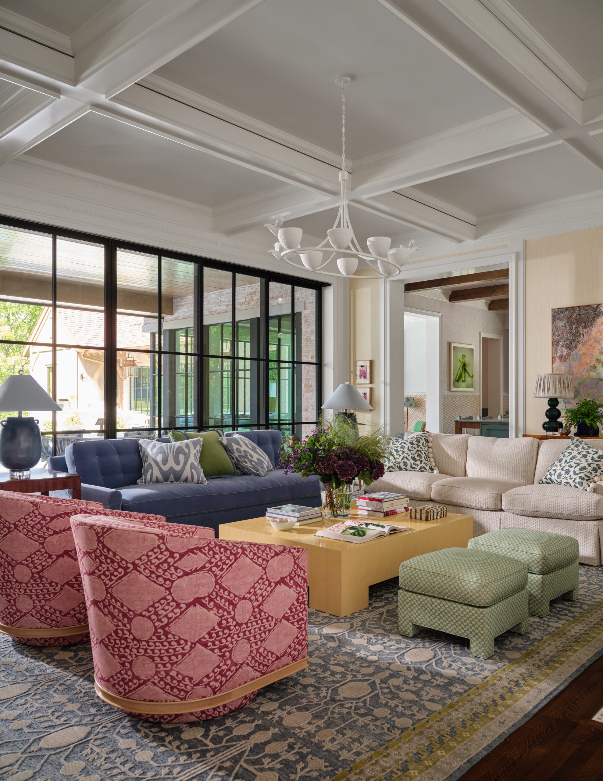 Living room featuring a coffered ceiling, blue sofa, beige sofa, pink patterned swivel chairs, green ottomans, a modern rectangular coffee table, and an ornate white chandelier designed by Collins Interiors.
