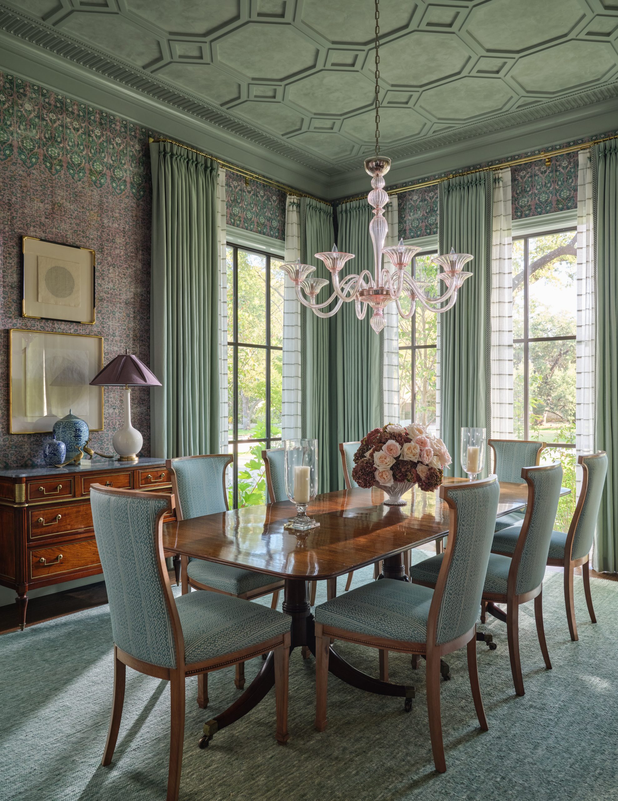 Dining room featuring a green geometric coffered ceiling, green curtains, floral wallpaper, a polished wood dining table with blue upholstered chairs, a vintage wood buffet, and a pink glass chandelier designed by Collins Interiors.
