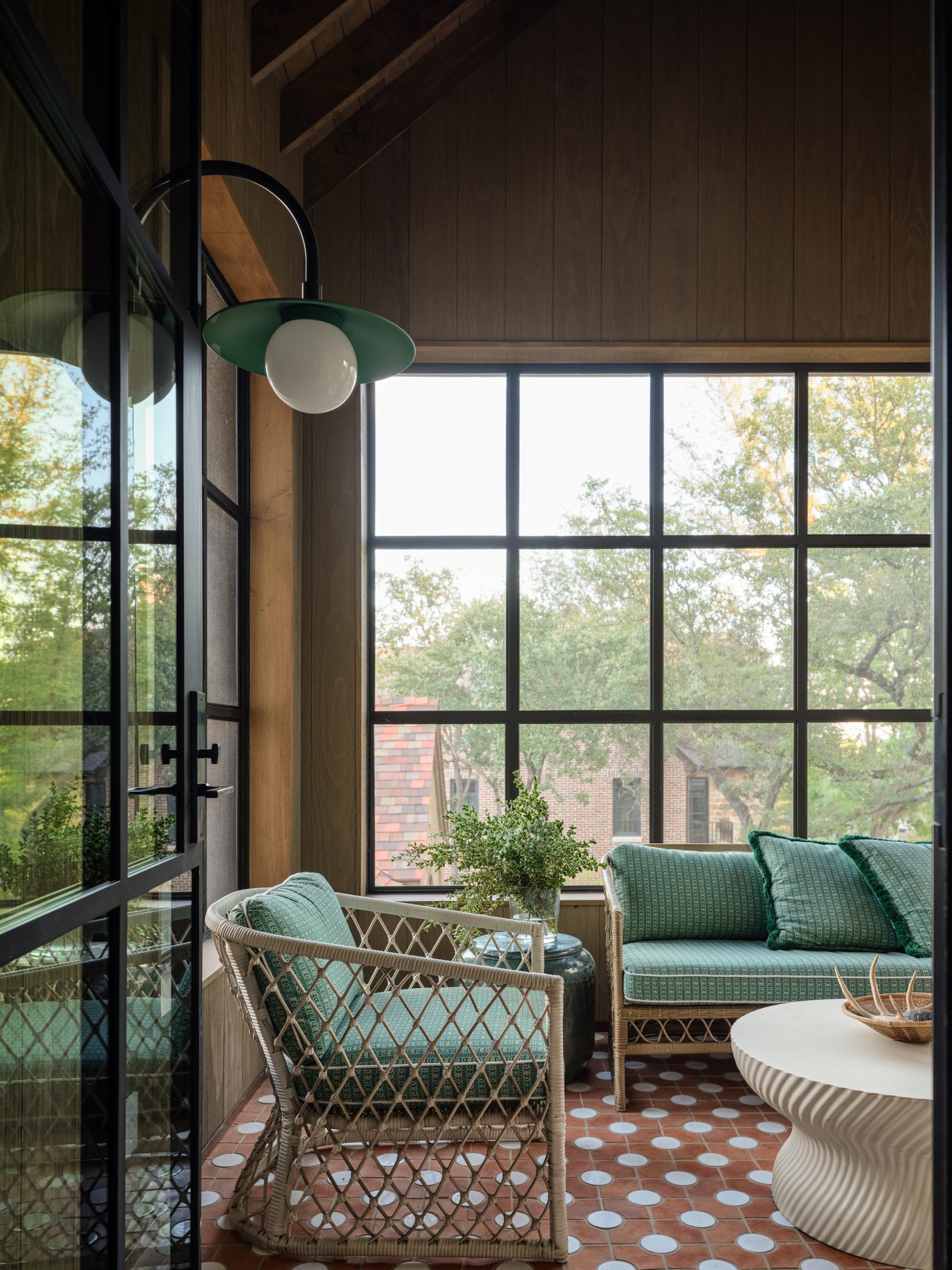 Sunroom porch featuring wood paneled walls and ceiling, large grid windows, green wicker outdoor furniture, terracotta patterned tile flooring, a modern white fluted coffee table, and a green gooseneck wall sconce designed by Collins Interiors.
