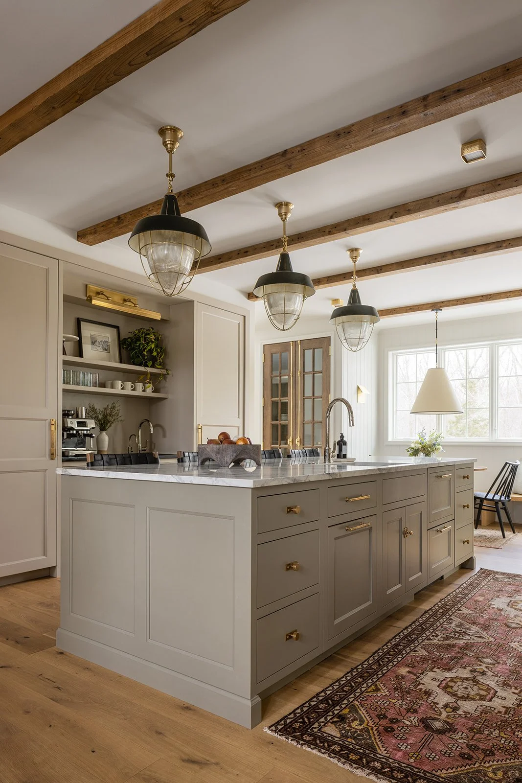 Luxury transitional kitchen featuring exposed ceiling wood beams, a light gray central island, an antique pattern runner rug, and custom glass-front pantry doors.
