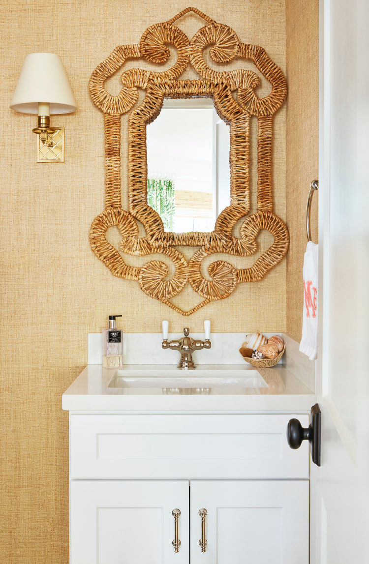 Charming coastal powder room featuring a white vanity with brass hardware, textured natural grasscloth wallpaper, and a statement ornate woven rattan scrollwork mirror