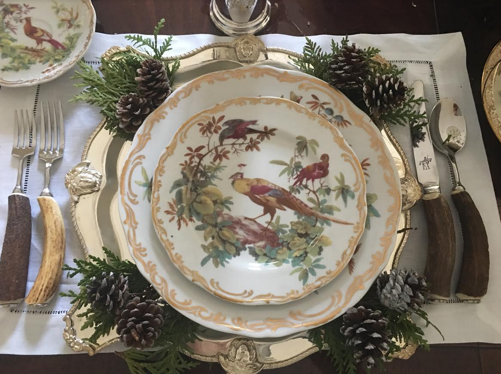 A sophisticated Thanksgiving tablescape featuring bird-patterned plates stacked on large silver chargers, paired with rustic antler flatware, fresh cedar sprigs, and small pinecones at each place setting.