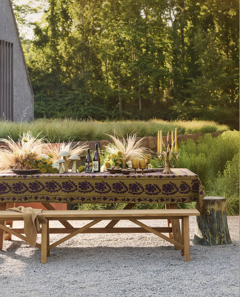An elegant outdoor Thanksgiving tablescape featuring a burgundy and gold tablecloth draped over a wooden table with a bench, beautifully styled with candles and natural grasses.