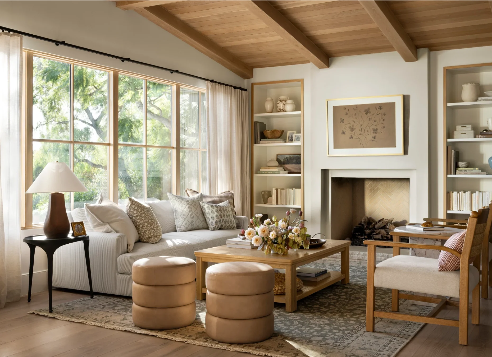 Warm transitional living room featuring a vaulted wood-beam ceiling, built-in bookshelves flanking a plaster fireplace, a neutral sofa with patterned pillows, and round leather pouf stools