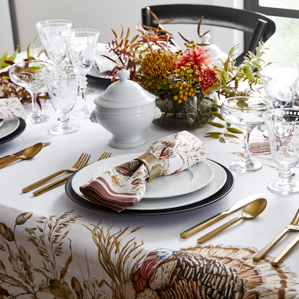 An elegant Thanksgiving table setting featuring crystal glassware, gold flatware, and white china plates juxtaposed against a whimsical turkey tablecloth and a natural centerpiece."